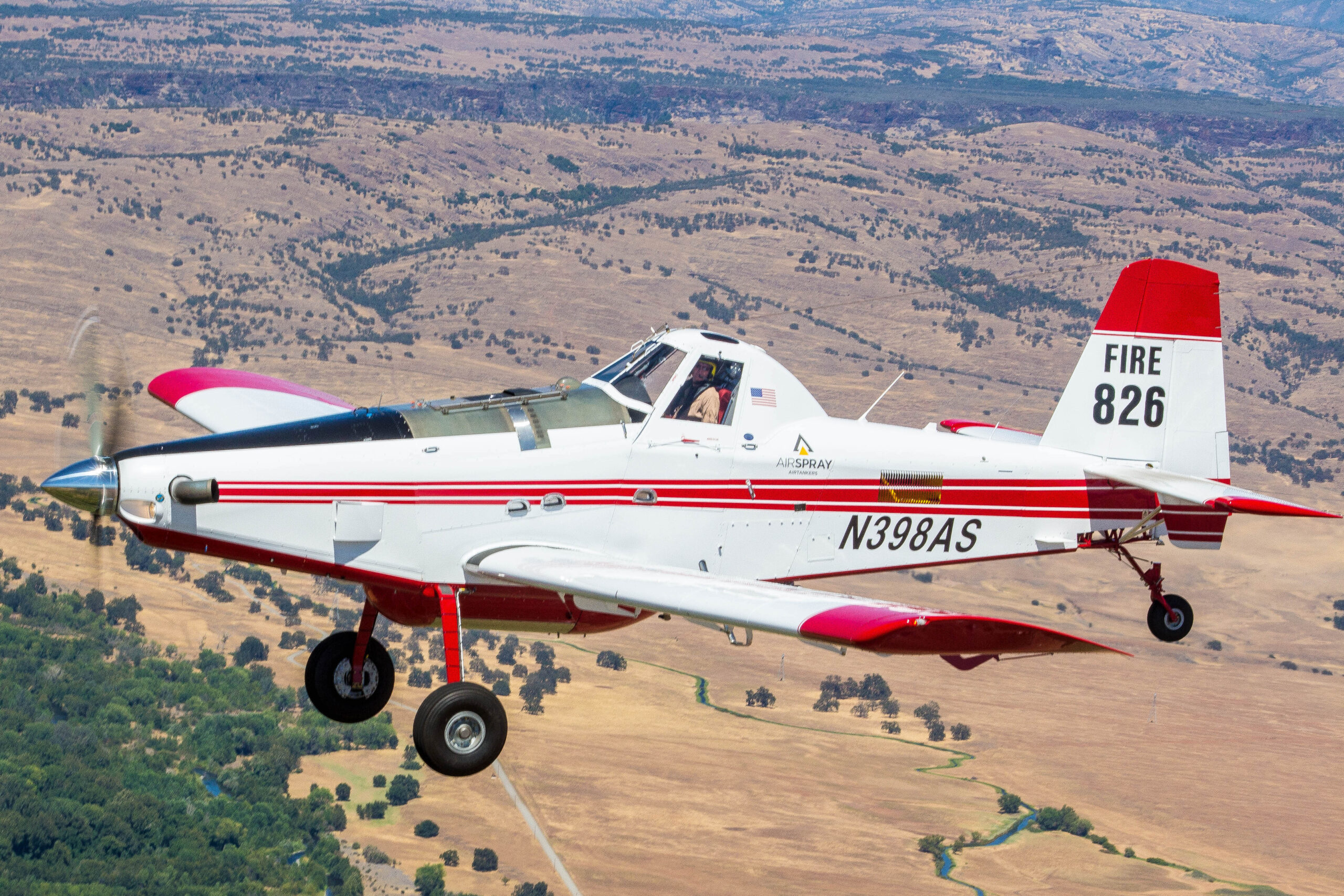 Air Tractor AT-802f firefighting aircraft owned by AirSpray in flight, equipped for aerial firefighting operations. The plane features a red and white color scheme, with registration number N398AS and designation "FIRE 826" on the tail. The background showcases a landscape suitable for fire suppression activities.