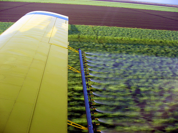 Aerial view of an Air Tractor crop duster in action, showcasing the wing and spray nozzles as it applies agricultural chemicals over a field of crops.