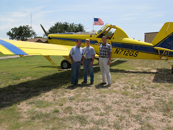 Three men stand beside an Air Tractor AT-502B agricultural aircraft, showcasing its capabilities in crop dusting and aerial application services. The plane, identified by the registration number N712GS, is positioned on a grassy field with a clear sky in the background.