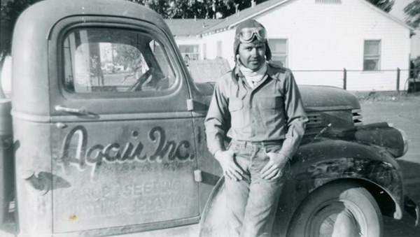 Historical photograph of a pilot standing beside a vintage truck with "Agai Inc." branding, showcasing early agricultural aviation practices. The pilot is wearing goggles and a flight suit, reflecting the era of crop dusting and aerial application.
