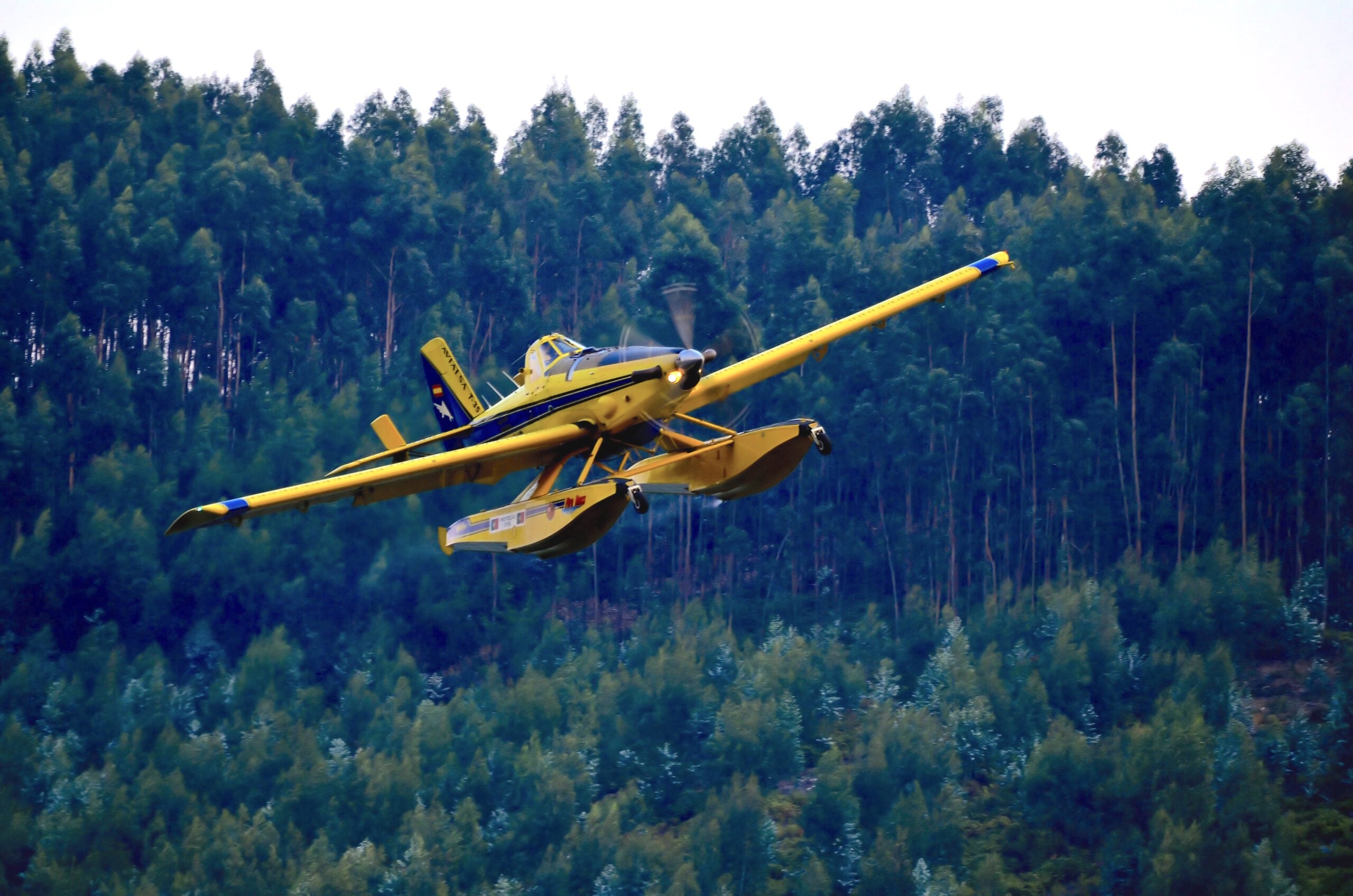 Air Tractor AT-802A agricultural aircraft in flight, featuring floatation gear for water operations, with a backdrop of dense forest. Ideal for aerial application and crop management.