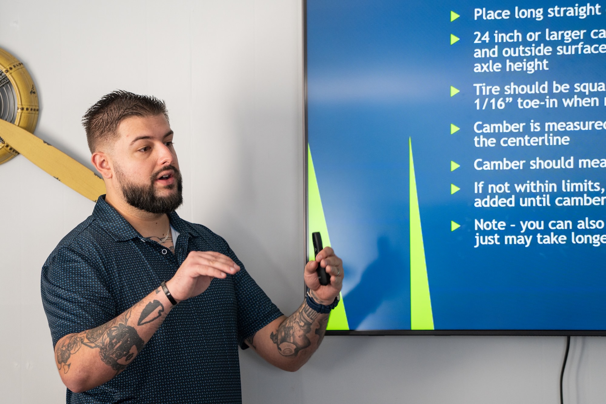 A man presents maintenance tips for agricultural aircraft, focusing on tire alignment and camber adjustments, with a visual aid in the background. The discussion may relate to models like the Air Tractor AT-502 or AT-802, known for their performance in crop dusting and agricultural applications.