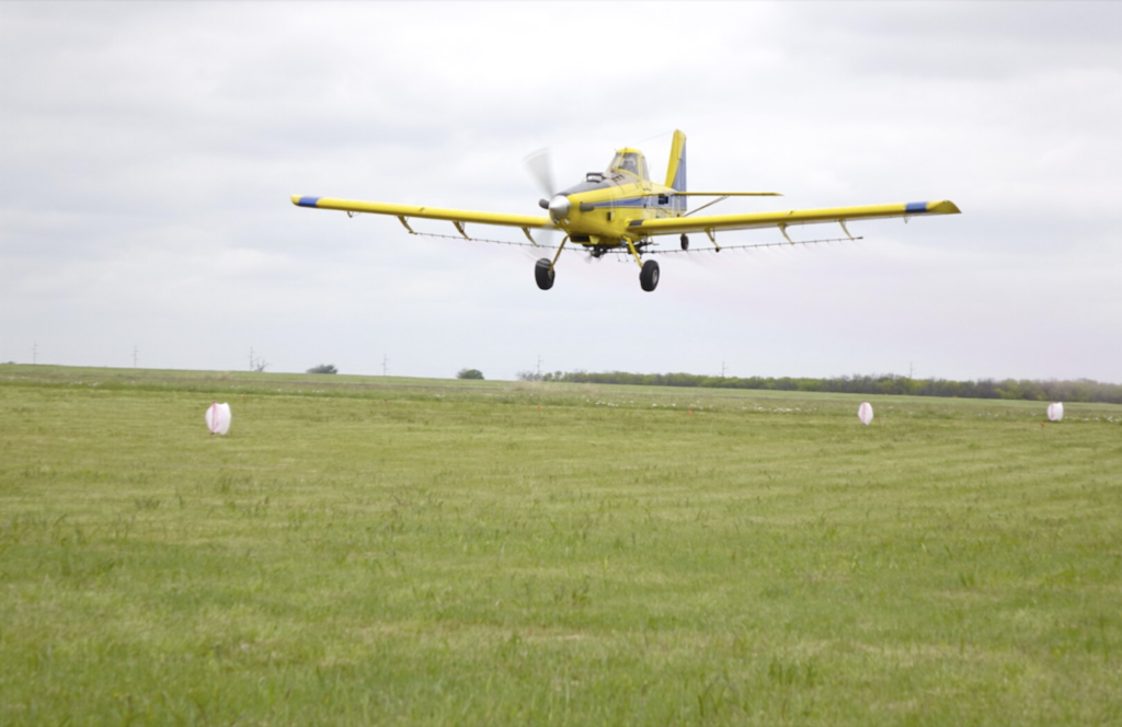 Air Tractor AT-502B agricultural aircraft flying low over a grassy field, demonstrating aerial application capabilities. This photo illustrates an article about Air Tractor hosting Operation Fly-In in Olney, Texas.
