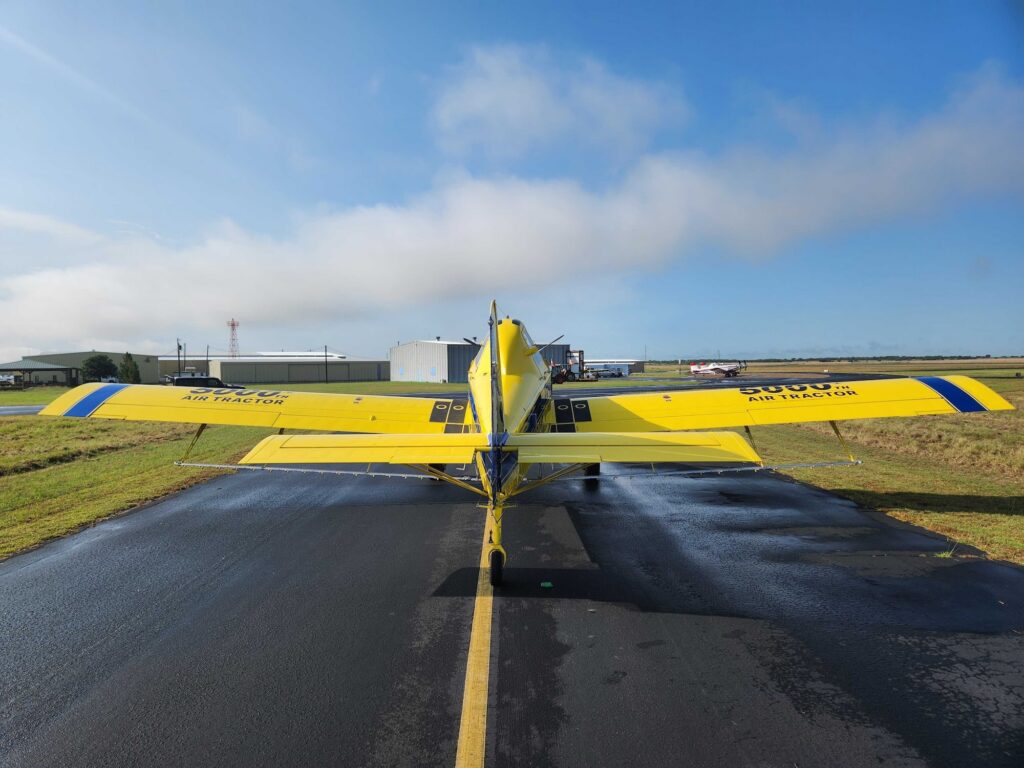 Air Tractor AT-502B agricultural aircraft parked on a runway, showcasing its distinctive design and operational capabilities. The background features a hangar and additional aircraft, highlighting its use in farming and aerial application.